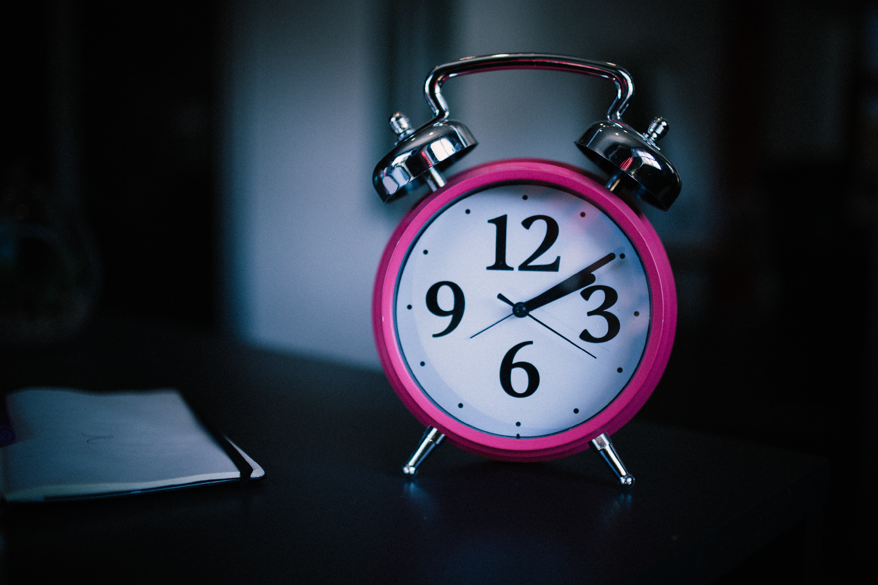 Em uma mesa de cabeceira há um relógio despertador rosa, com os números 3, 6, 9 e 12
