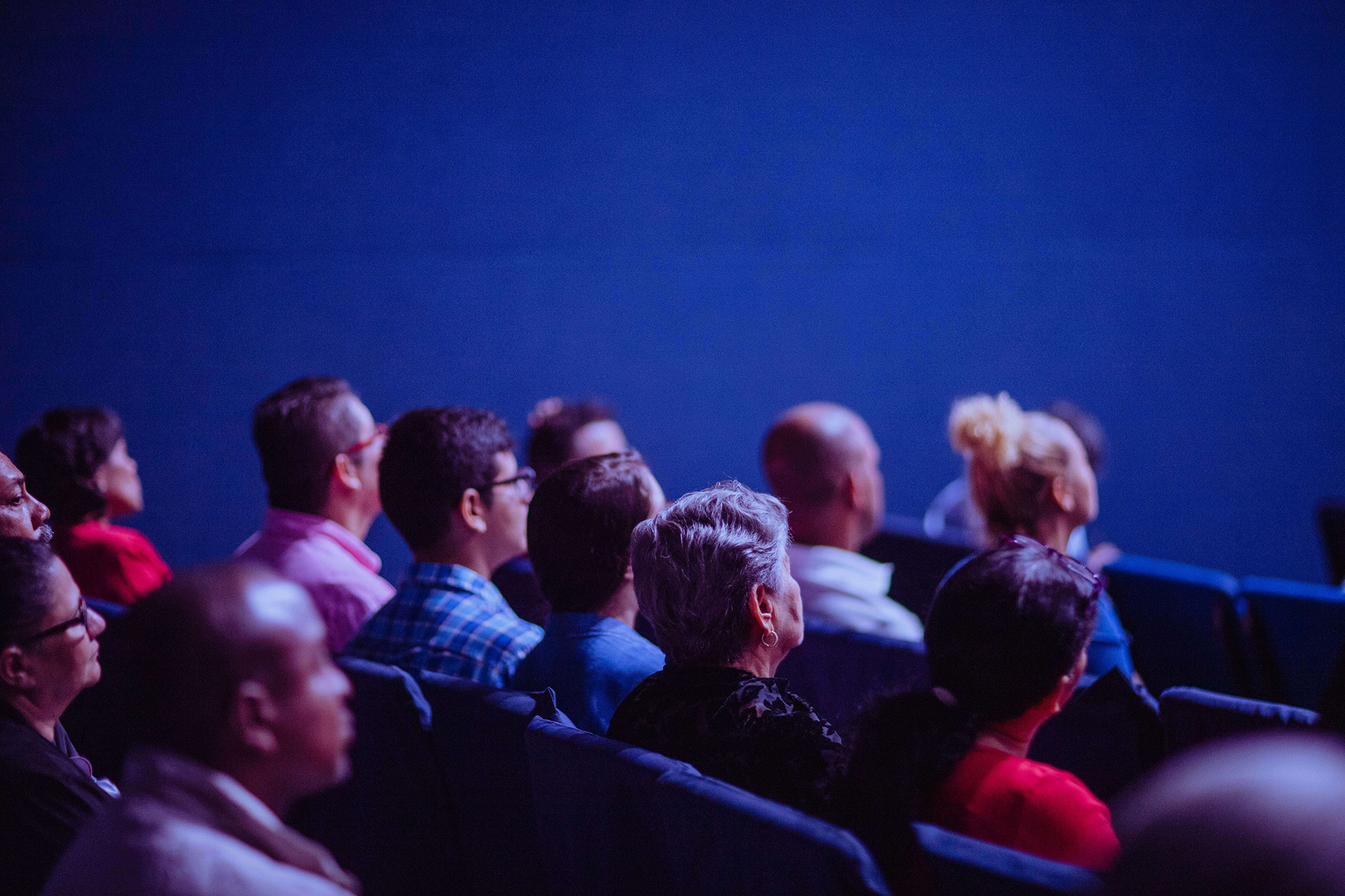 É um auditório de fundo azul, com diversas pessoas sentadas em cadeiras assistindo à alguma palestra.